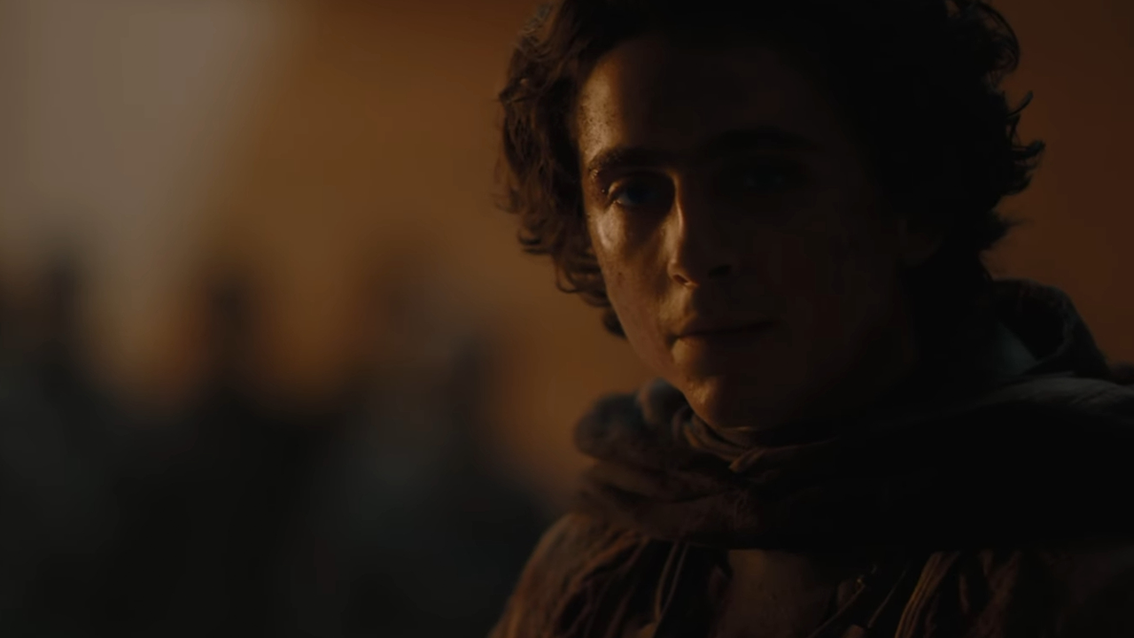 El actor Timothée Chalamet, en el personaje de Paul Atreides, en primer plano, mirando hacia adelante contra un fondo desértico borroso con varias personas en el fondo, al anochecer, representando al elenco de Dune 2.