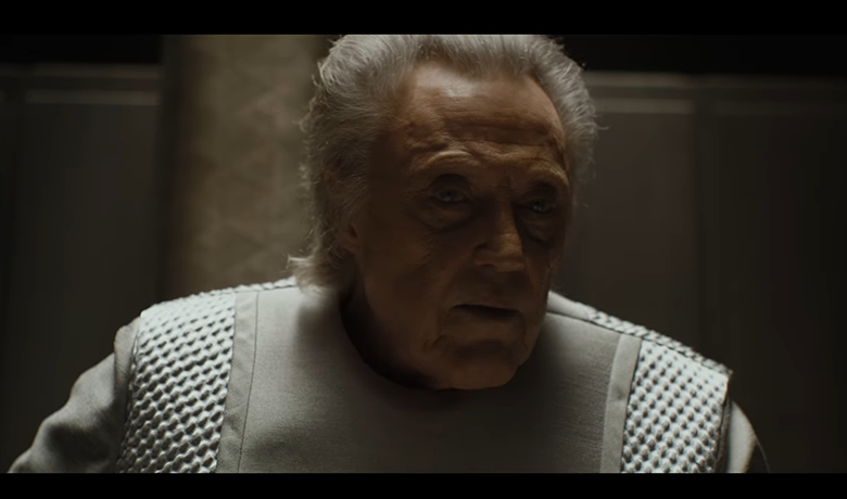 El actor Christopher Walken, retratado como un hombre mayor, de cabello gris y mirada perdida, en primer plano, vestido con una túnica gris con detalles plateados, sentado en un trono gris, en un ambiente oscuro, con una luz fuerte y concentrada que viene desde arriba.