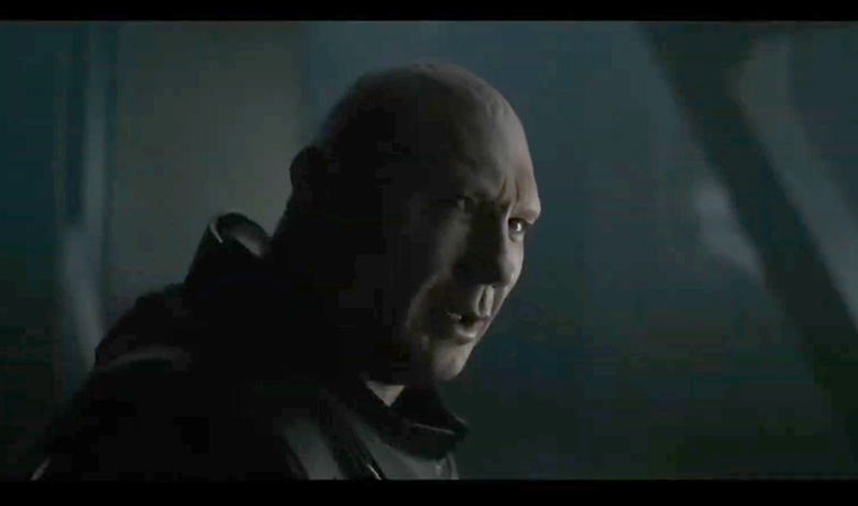 El actor Dave Bautista es representado como un hombre corpulento con una armadura oscura, con piel muy pálida y sin pelo en la cara ni en las cejas, en un primer plano, mirando hacia la derecha en las sombras.