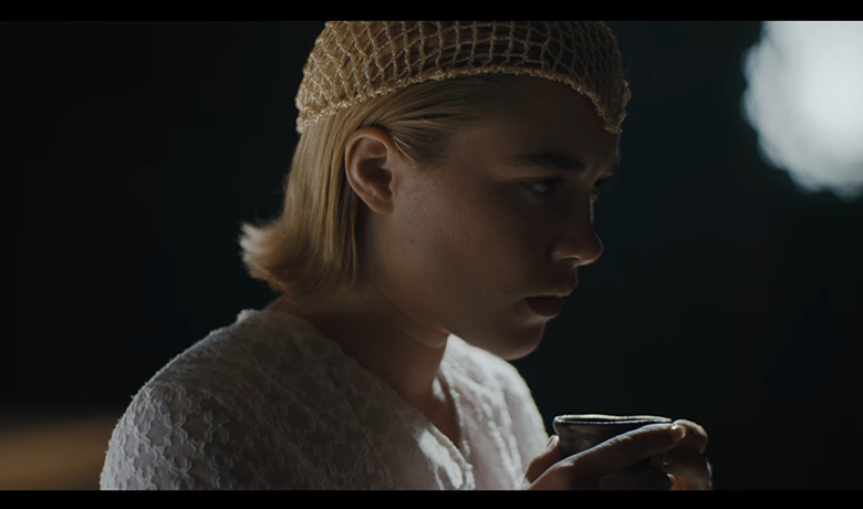 La actriz Florence Pugh es representada como una mujer con cabello rubio cortado por encima de los hombros, luciendo un sombrero hecho con hilos dorados y un vestido blanco, en un entorno muy oscuro pero con una fuerte luz brillando sobre ella.