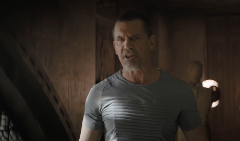 El actor Josh Brolin, luciendo un aspecto militar agresivo con una perilla gris y cabello corto y negro, viste una camiseta gris con rayas en el pecho y mira hacia adelante en un entorno oscuro en tonos madera.