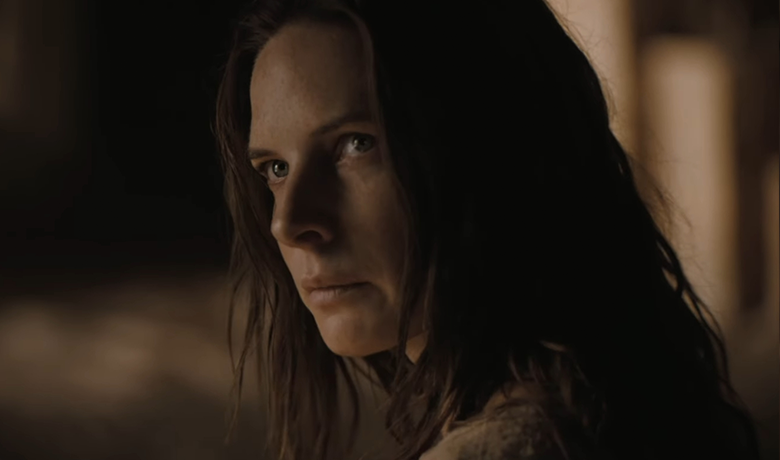 La actriz Rebecca Ferguson, retratada como una mujer de cabello oscuro y despeinado, en primer plano, mirando hacia la izquierda con una mirada fría y agresiva, con un vestido marrón, en un entorno poco iluminado.