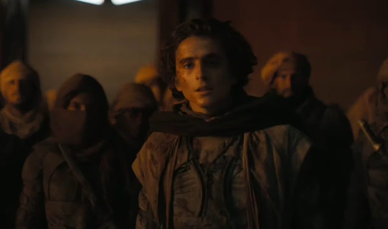 El actor Timothée Chalamet, representado como un joven de pelo despeinado, vestido con ropa marrón y un pañuelo oscuro sobre los hombros, aparece en un primer plano, mirando hacia delante contra un fondo desértico borroso con varias personas en el fondo al anochecer.