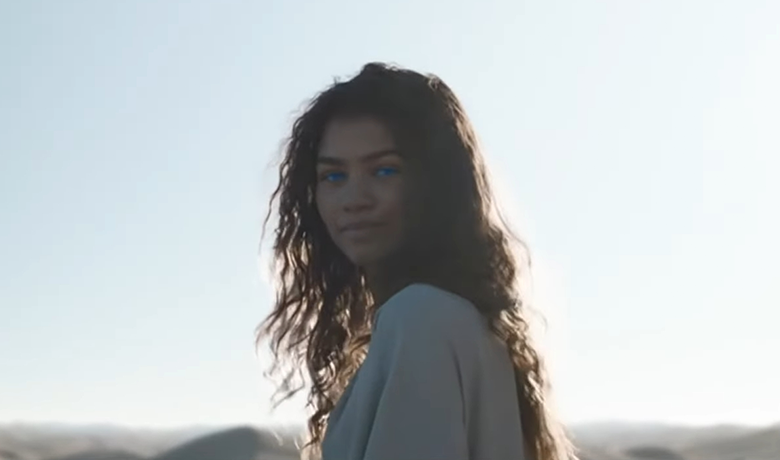 La actriz Zendaya es representada como una mujer de cabello largo, oscuro y desordenado, ojos azules y un vestido blanco. El escenario es el desierto al mediodía, con dunas borrosas al fondo.
